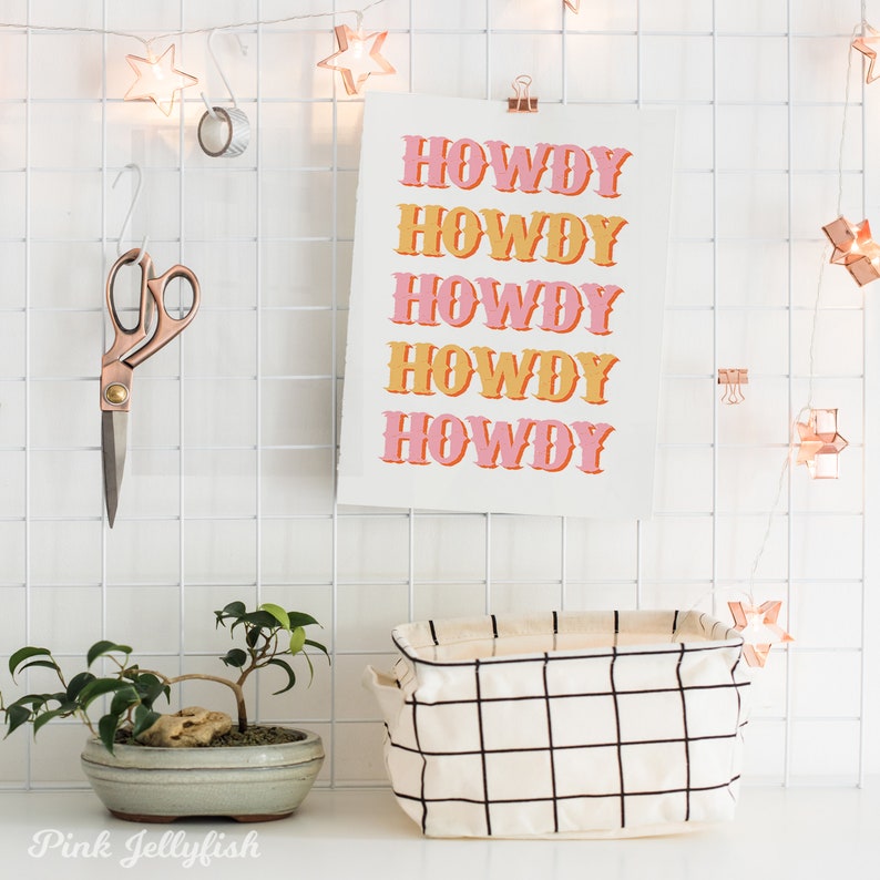 May include: A white grid wall with a pink and orange print that says "Howdy" five times. There is a small potted bonsai tree, a white and black grid storage basket, and a pair of scissors hanging on the wall.