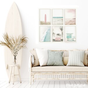 May include: A set of six framed prints with a beach theme. The prints feature a pink surfboard, a beach scene with waves, a pink car, and a lifeguard stand. The prints are arranged in a grid pattern on a white wall.