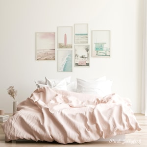 May include: A pink bed with white pillows and a white wall with six framed pictures of beach scenes. The pictures include a pink surfboard, a beach with people walking, a beach with a lifeguard stand, a beach with a car, and a beach with waves.