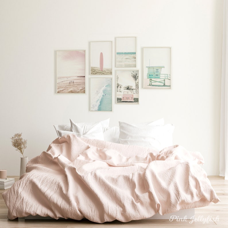 May include: A pink bed with white pillows and a white wall with six framed pictures of beach scenes. The pictures include a pink surfboard, a beach with people walking, a beach with a lifeguard stand, a beach with a car, and a beach with waves.