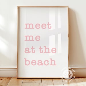 Puede incluir: Una impresi&oacute;n enmarcada con las palabras "meet me at the beach" en letras rosas sobre un fondo blanco. La impresi&oacute;n est&aacute; en un marco de madera clara y se muestra sobre una superficie de madera.