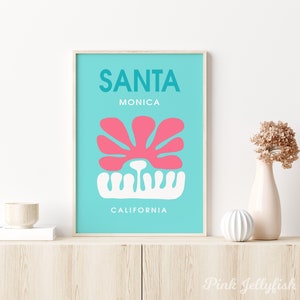 May include: A turquoise blue poster with the text "SANTA MONICA CALIFORNIA" in white lettering. A pink stylized flower with a white stem is in the center of the poster.