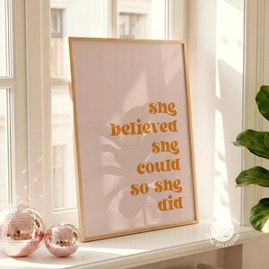 Op de afbeelding: Een ingelijste print met de tekst "She believed she could so she did" in oranje letters op een roze achtergrond. De print zit in een licht houten frame. Twee decoratieve discoballen staan op de voorgrond.