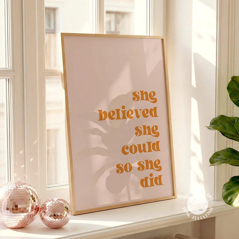Op de afbeelding: Een ingelijste print met de tekst "She believed she could so she did" in oranje letters op een roze achtergrond. De print zit in een licht houten frame. Twee decoratieve discoballen staan op de voorgrond.