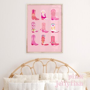 May include: A framed print featuring nine colorful cowboy boots in shades of pink, orange, and white, set against a pink background. The boots showcase various designs, including floral patterns and cactus motifs. The print is displayed above a bed with a woven headboard.