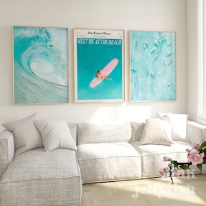 May include: Three framed prints with a beach theme. The left print shows a blue wave crashing in the ocean. The middle print features a woman on a pink surfboard in the ocean with the text "The Coastal Times" and "Meet Me At The Beach". The right print shows a top-down view of the ocean with several people on surfboards.