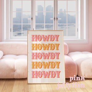 May include: A framed print with the word "Howdy" repeated five times in a stacked vertical design. The word is in a pink and orange color scheme with a retro font style. The print is on a white background and has a gold frame.