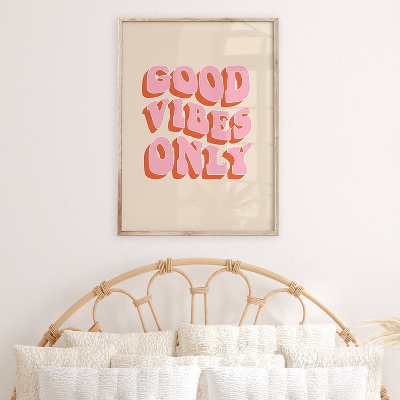 May include: A framed print with a beige background and the text "GOOD VIBES ONLY" in pink and orange letters. The print is hanging on a white wall above a wicker headboard.