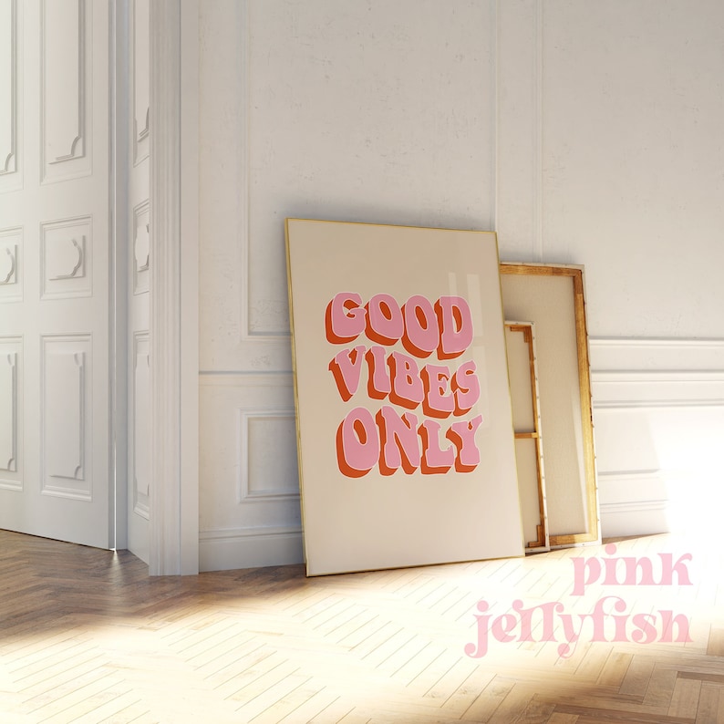 May include: A framed print with a beige background and the text "GOOD VIBES ONLY" in pink and orange letters. The print is in a gold frame and is leaning against a white wall.