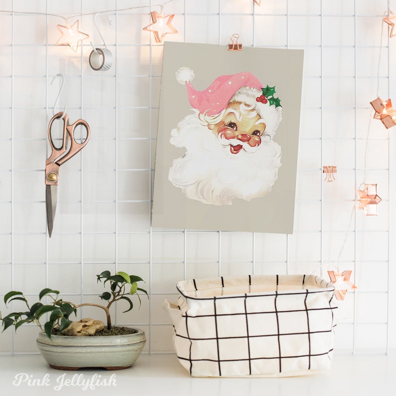 May include: A vintage illustration of Santa Claus's face with a pink hat and white beard, printed on a light grey background. The illustration is framed and hanging on a white grid wall with string lights.
