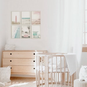 May include: A nursery room with a white crib, a dresser, and a white basket. The walls are white and there are six framed prints of beach scenes on the wall. The prints feature a surfboard, a beach scene, a lifeguard stand, and a vintage car.