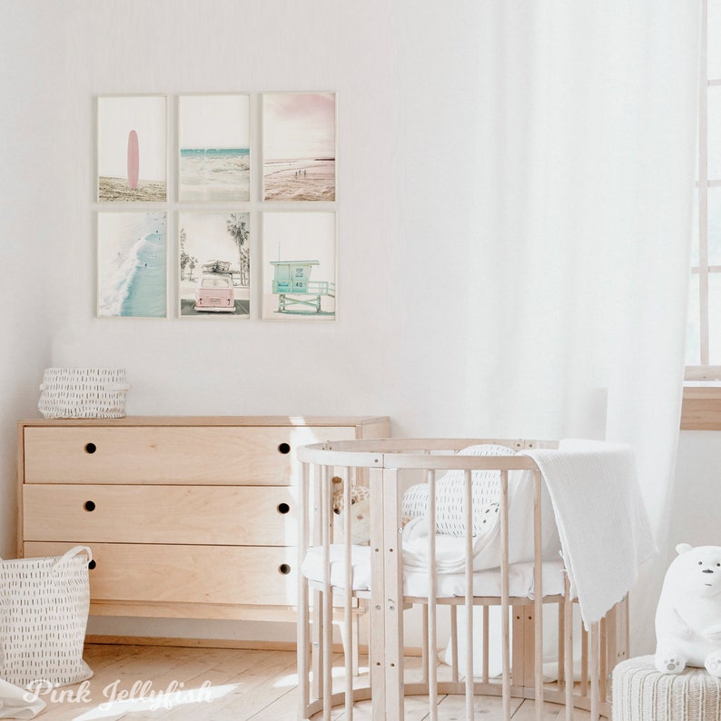 May include: A nursery room with a white crib, a dresser, and a white basket. The walls are white and there are six framed prints of beach scenes on the wall. The prints feature a surfboard, a beach scene, a lifeguard stand, and a vintage car.