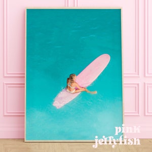 May include: A woman sits on a pink surfboard in turquoise water. The surfboard is long and narrow, and the woman is wearing a white bikini. The water is clear and blue, and the sky is a bright blue.