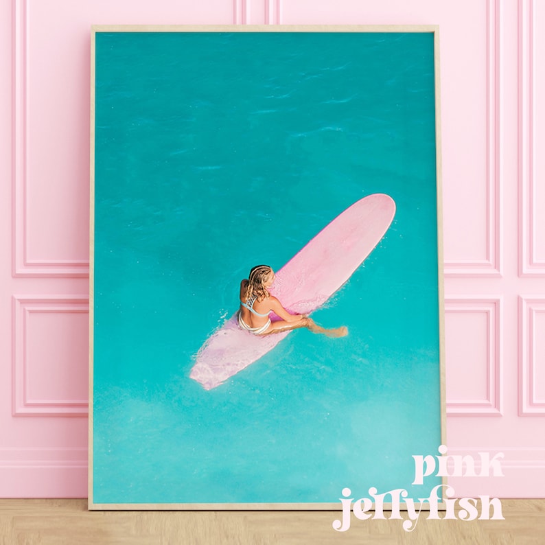 May include: A woman sits on a pink surfboard in turquoise water. The surfboard is long and narrow, and the woman is wearing a white bikini. The water is clear and blue, and the sky is a bright blue.