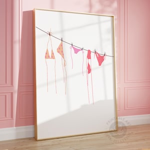 Puede incluir: Una impresi&oacute;n enmarcada que presenta una ilustraci&oacute;n minimalista de varios bikinis colgados de un tendedero. Los bikinis son en tonos rosa y melocot&oacute;n, con varios patrones. El fondo es blanco y el marco es dorado.