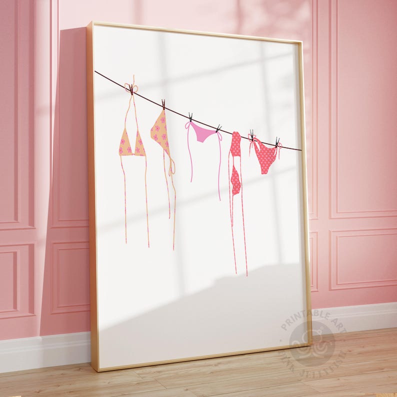 Puede incluir: Una impresi&oacute;n enmarcada que presenta una ilustraci&oacute;n minimalista de varios bikinis colgados de un tendedero. Los bikinis son en tonos rosa y melocot&oacute;n, con varios patrones. El fondo es blanco y el marco es dorado.