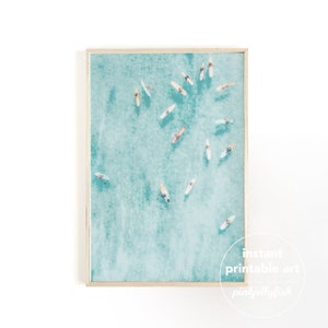 May include: An aerial view of a turquoise ocean with multiple surfers paddling on their surfboards. The image is framed in a light wood frame.