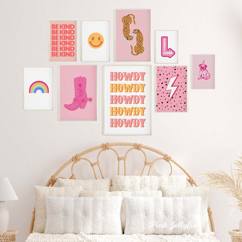 Pode incluir: Uma parede de galeria com impress&otilde;es emolduradas em rosa e branco que apresentam um arco-&iacute;ris, uma bota de cowboy, um rosto sorridente, estampa de leopardo, um raio e as palavras "Howdy" repetidas cinco vezes. As impress&otilde;es est&atilde;o penduradas em uma parede branca acima de uma cabeceira de vime.