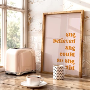 Op de afbeelding: Een roze retro-stijl broodrooster met chromen accenten staat naast een ingelijste print met de tekst "She believed she could so she did". Een witte theekop en een zwart-wit zigzagpatroon mok staan ook op het houten oppervlak.