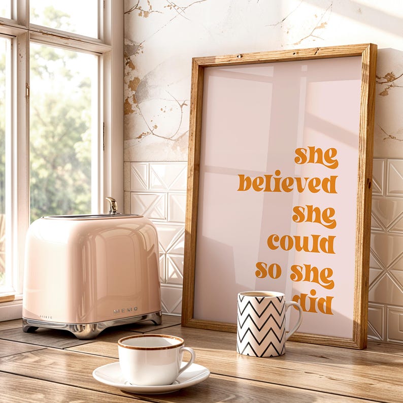 Op de afbeelding: Een roze retro-stijl broodrooster met chromen accenten staat naast een ingelijste print met de tekst "She believed she could so she did". Een witte theekop en een zwart-wit zigzagpatroon mok staan ook op het houten oppervlak.