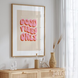 May include: A framed print with a light brown background and pink text that reads "GOOD VIBES ONLY" in a retro font style. The print is hanging on a white wall above a wooden dresser with a wicker vase and a bottle.