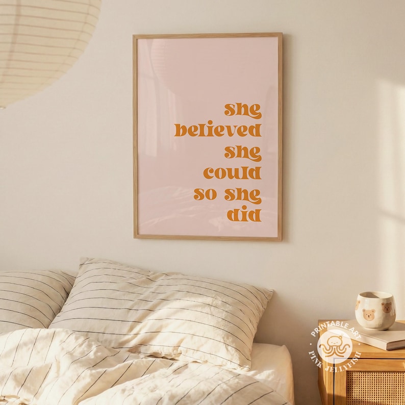 Op de afbeelding: Een ingelijste print met de tekst "She believed she could so she did" in oranje letters op een roze achtergrond. De print staat in een licht houten lijst en wordt boven een bed weergegeven.