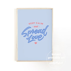 May include: A framed print with a pale blue background. The text "KEEP CALM - and - Spread Love" is written in blue script with a red heart. The frame is a light wood colour. The words "instant printable art" and "pinkjellyfish" are in the bottom right corner.