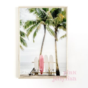 May include: A framed print of a beach scene with three surfboards leaning against a palm tree. The surfboards are white with a pink surfboard in the middle.