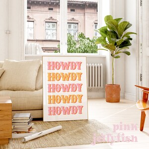 May include: A white poster with the word "HOWDY" repeated five times in a retro font. The word is in pink and orange, with a slight shadow effect. The poster is hanging on a wall in a living room setting.