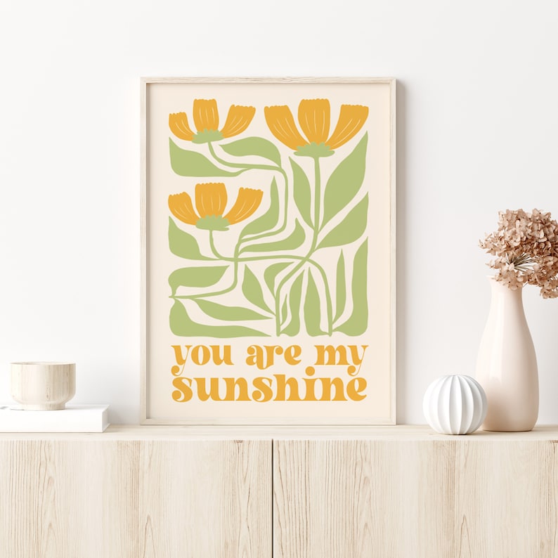 May include: A framed art print featuring a floral design with orange flowers and green leaves on a cream background. The text "you are my sunshine" is in orange. The print is displayed on a light wood cabinet.