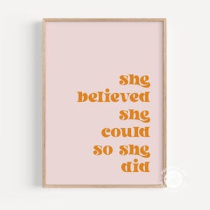 Op de afbeelding: Een ingelijste kunstdruk met een lichtroze achtergrond en de woorden "she believed she could so she did" in oranje letters. De print zit in een lichtbruine houten lijst.