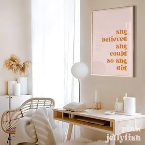 Op de afbeelding: Een ingelijste print met de tekst "she believed she could so she did" in oranje letters. De print hangt op een lichtroze achtergrond met een gouden lijst. De kamer heeft een bureau, stoel en decoratieve items.
