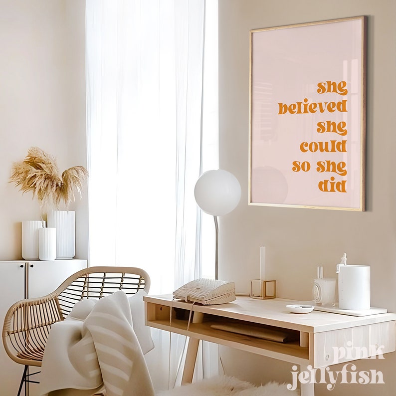 Op de afbeelding: Een ingelijste print met de tekst "she believed she could so she did" in oranje letters. De print hangt op een lichtroze achtergrond met een gouden lijst. De kamer heeft een bureau, stoel en decoratieve items.