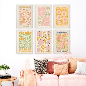 May include: Six framed floral art prints with the words "Flower Market" and city names. The prints feature colorful botanical illustrations in pink, yellow, and green. A pink sofa with pillows and a blanket is in the foreground.