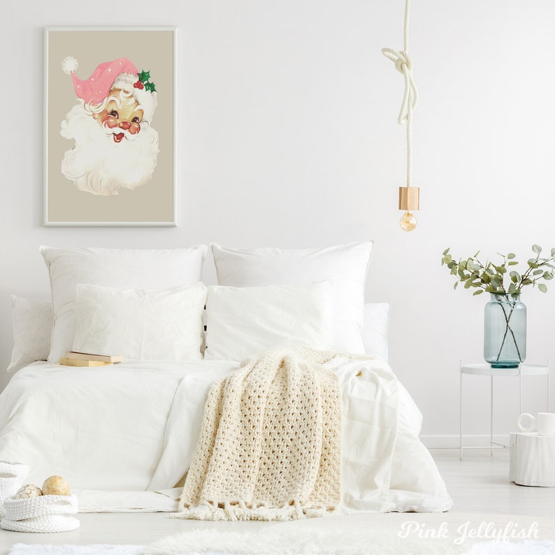 May include: A white bed with a white crocheted throw blanket and a framed print of a vintage Santa Claus illustration with a pink hat. The bed is styled with white pillows and a book on the bed. There is a small white table with a vase of green leaves and a white mug on it.