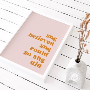Op de afbeelding: Een ingelijste print met de tekst "she believed she could so she did" in oranje letters op een roze achtergrond. De print zit in een witte lijst en staat naast een witte vaas met gedroogde bloemen.