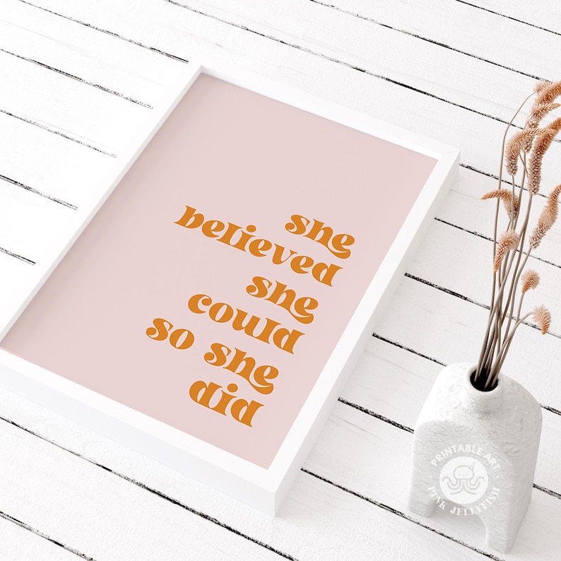 Op de afbeelding: Een ingelijste print met de tekst "she believed she could so she did" in oranje letters op een roze achtergrond. De print zit in een witte lijst en staat naast een witte vaas met gedroogde bloemen.