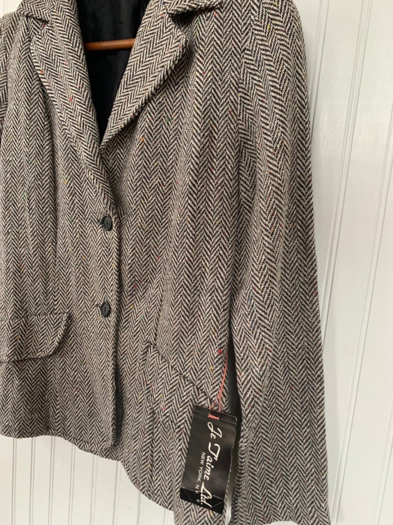 Op de afbeelding: Een zwart-witte herringbone blazer met een enkele knoopsluiting en een zak aan de linkerkant. De blazer heeft een label met de tekst "Je Taime Ltd. New York, N.Y."