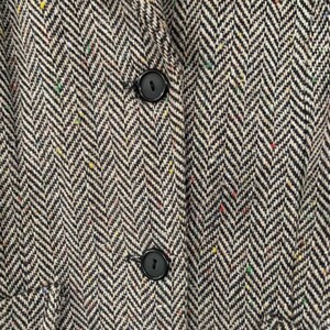 Op de afbeelding: Close-up van een zwart-witte herringbone tweed stof met twee zwarte knopen. De stof heeft een subtiel gespikkeld patroon.