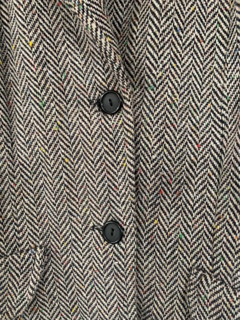 Op de afbeelding: Close-up van een zwart-witte herringbone tweed stof met twee zwarte knopen. De stof heeft een subtiel gespikkeld patroon.