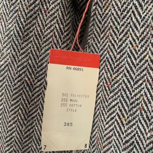 Op de afbeelding: Een wit label met zwarte tekst bevestigd aan een zwart-witte herringbone stof. Het label vermeldt "RN 46891", "50% Polyester", "25% Wol", "25% Katoen", "Style" en "385".