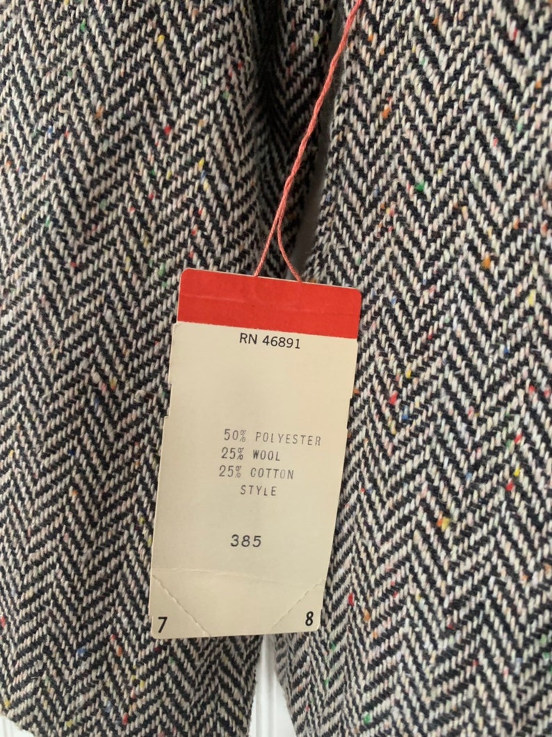 Op de afbeelding: Een wit label met zwarte tekst bevestigd aan een zwart-witte herringbone stof. Het label vermeldt "RN 46891", "50% Polyester", "25% Wol", "25% Katoen", "Style" en "385".