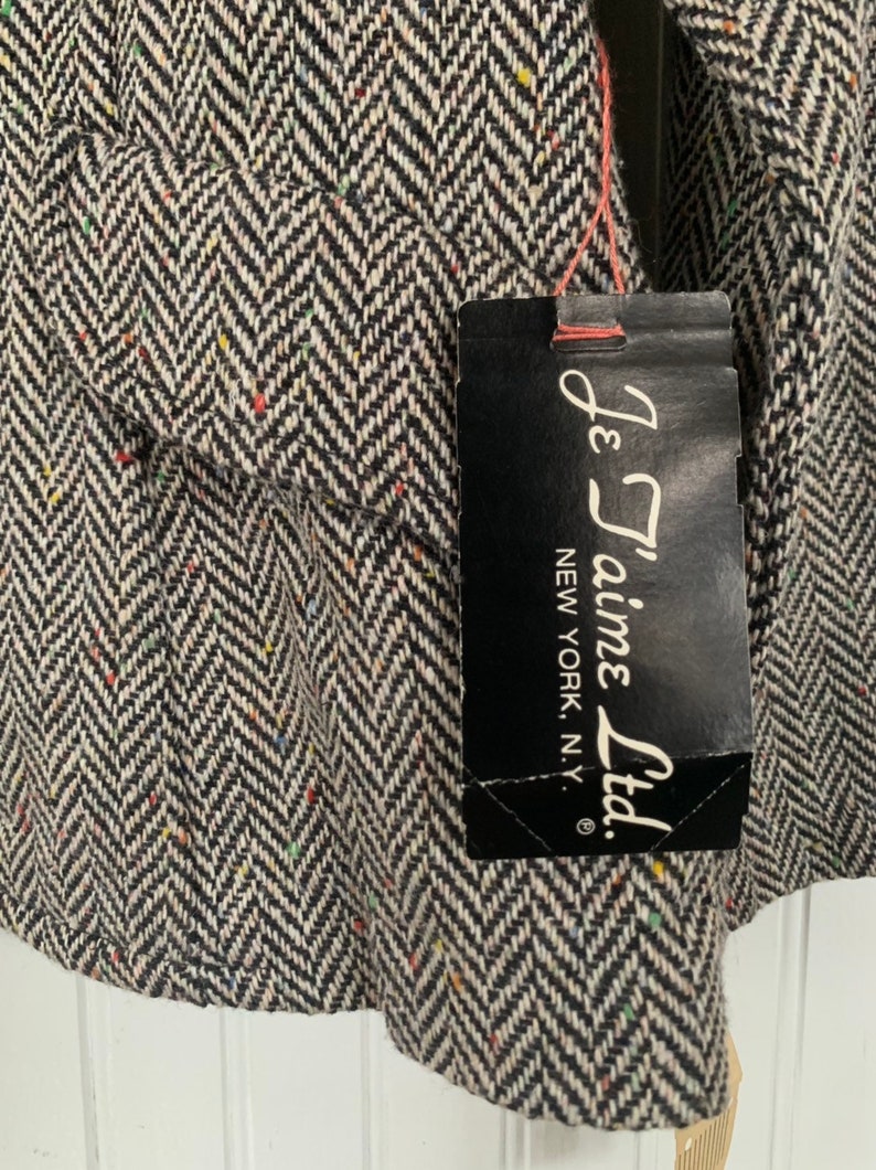 Op de afbeelding: Een zwart-witte herringbone tweed stof met een zwart label waarop "Je T'aime Ltd. New York, N.Y." staat.