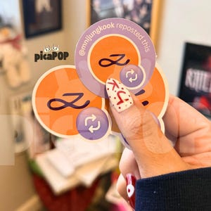 Puede incluir: Una colección de pegatinas redondas en naranja y morado. Las pegatinas presentan un símbolo de infinito estilizado y un icono de flecha circular. Una pegatina tiene el texto "@mnijungkook reposted this". Las pegatinas se sostienen en una mano.