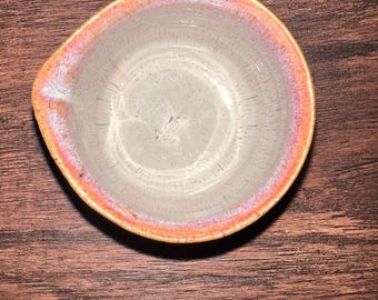 May include: A small, handmade ceramic bowl with a light gray interior and a rim that transitions from pink to orange. The bowl has a small spout and is set against a dark wood grain background.