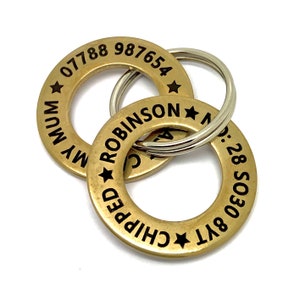 Op de afbeelding: Twee gouden metalen hondenpenningen met zwarte gegraveerde tekst. De bovenste penning heeft de tekst "MY MUM 07788 987654" en de onderste penning heeft de tekst "CHIPPED ROBINSON 877 5030 28."