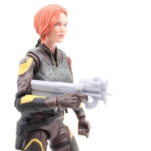 May include: A detailed action figure of a woman with red hair, wearing a brown and yellow tactical suit, holding a gray weapon. The figure has a serious expression and is set against a white background.