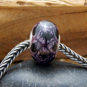 Könnte beinhalten: Eine dunkelviolette und schwarze Glasperle mit einem wirbelnden Muster. Die Perle befindet sich auf einer silbernen Kette.