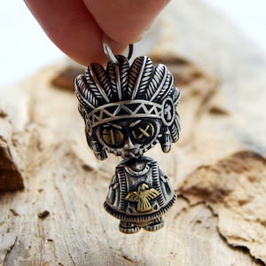 Puede incluir: Un colgante plateado con forma de muñeca nativa americana. La muñeca tiene un tocado de plumas, ojos redondos con pupilas en forma de cruz y un emblema de pájaro dorado en el pecho. El colgante está sujeto por una mano.