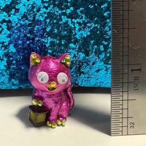 May include: A metallic magenta cat figurine with large white eyes, green glitter accents on the ears and paws, and a small brown lantern held in one paw. A metal ruler is positioned to the right, showing the figure is approximately 1.25 inches tall.